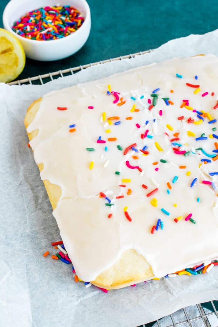 a square cake with icing and sprinkles on some parchment paper with a bowl of sprinkles in the background