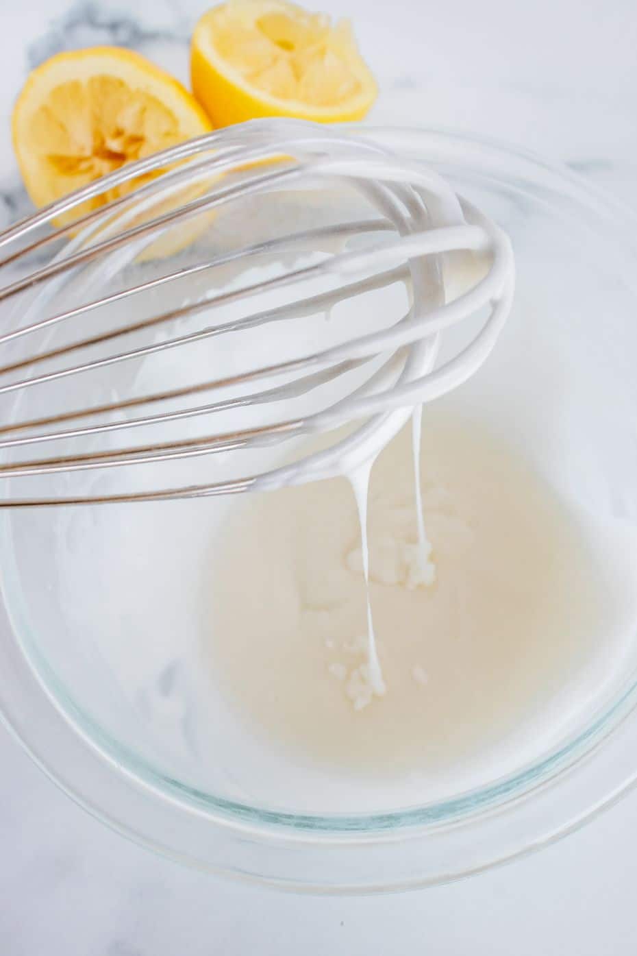 A whisk combing ingredients for a lemon glaze in a glass bowl with some squeezed lemons in the background