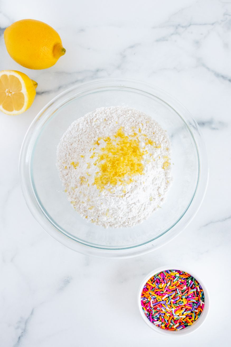 a clear bowl with flour, baking powder and lemon zest in it with a bowl of sprinkles and some chopped lemons in the background