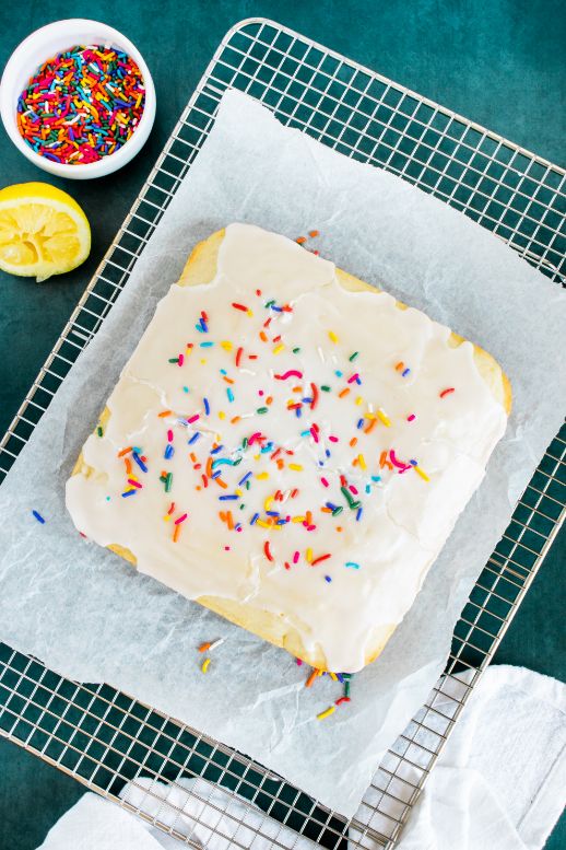 A cooled lemon cake on a wire rack with some parchment paper underneath it and a bowl of sprinkles on the side