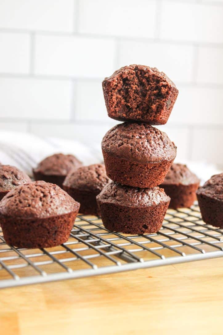 A stack of 3 chocolate muffins on a wire rack with a bite out of the muffin on top of the stack