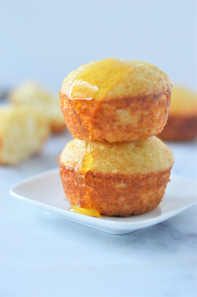 A stack of two golden muffins on a white plate with a drizzle of honey on top of the muffins and extra muffins in the background