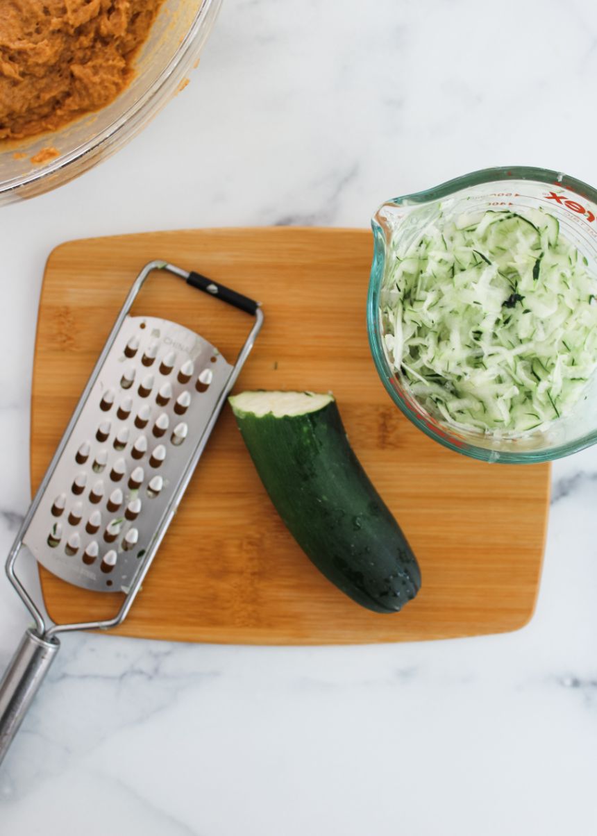 A zucchini being grated into a glass bowl