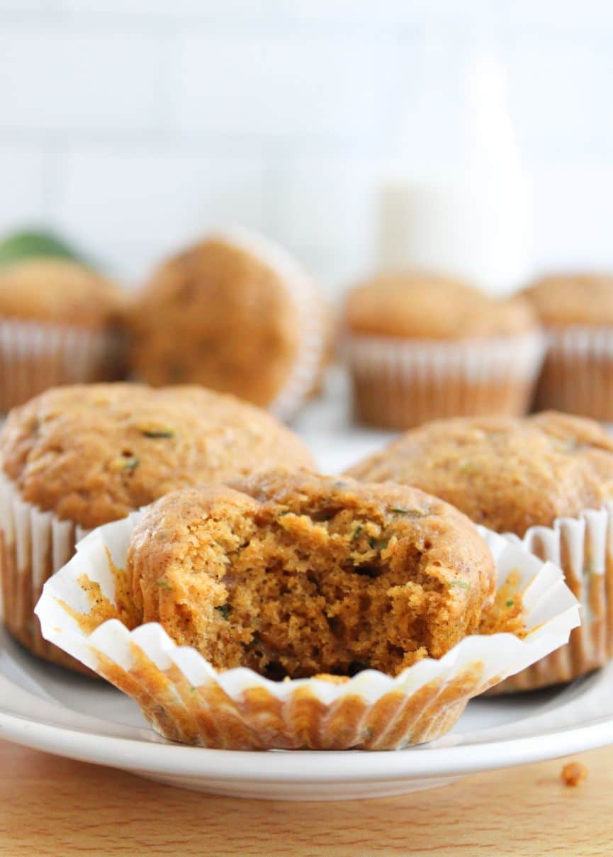 A pumpkin and zucchini muffin with a bite out of it served on a white plate with extra muffins in the background