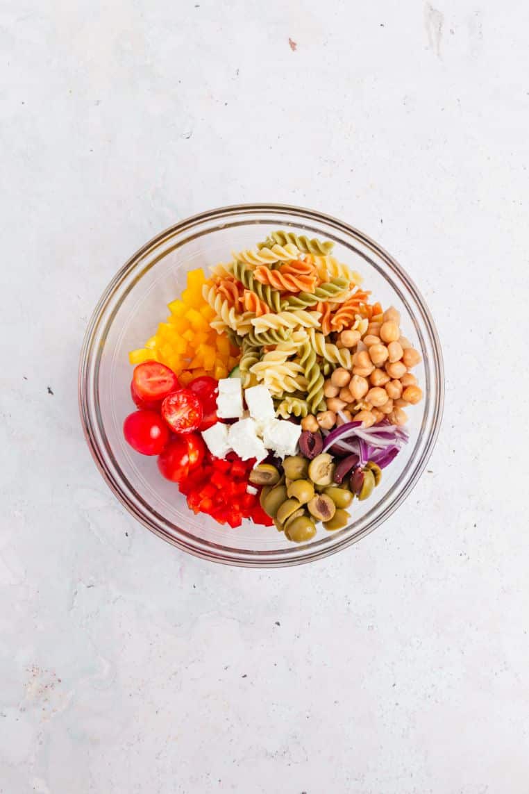 Overhead image of pasta, chickpeas, chopped veg and vinaigrette together in a glass bowl