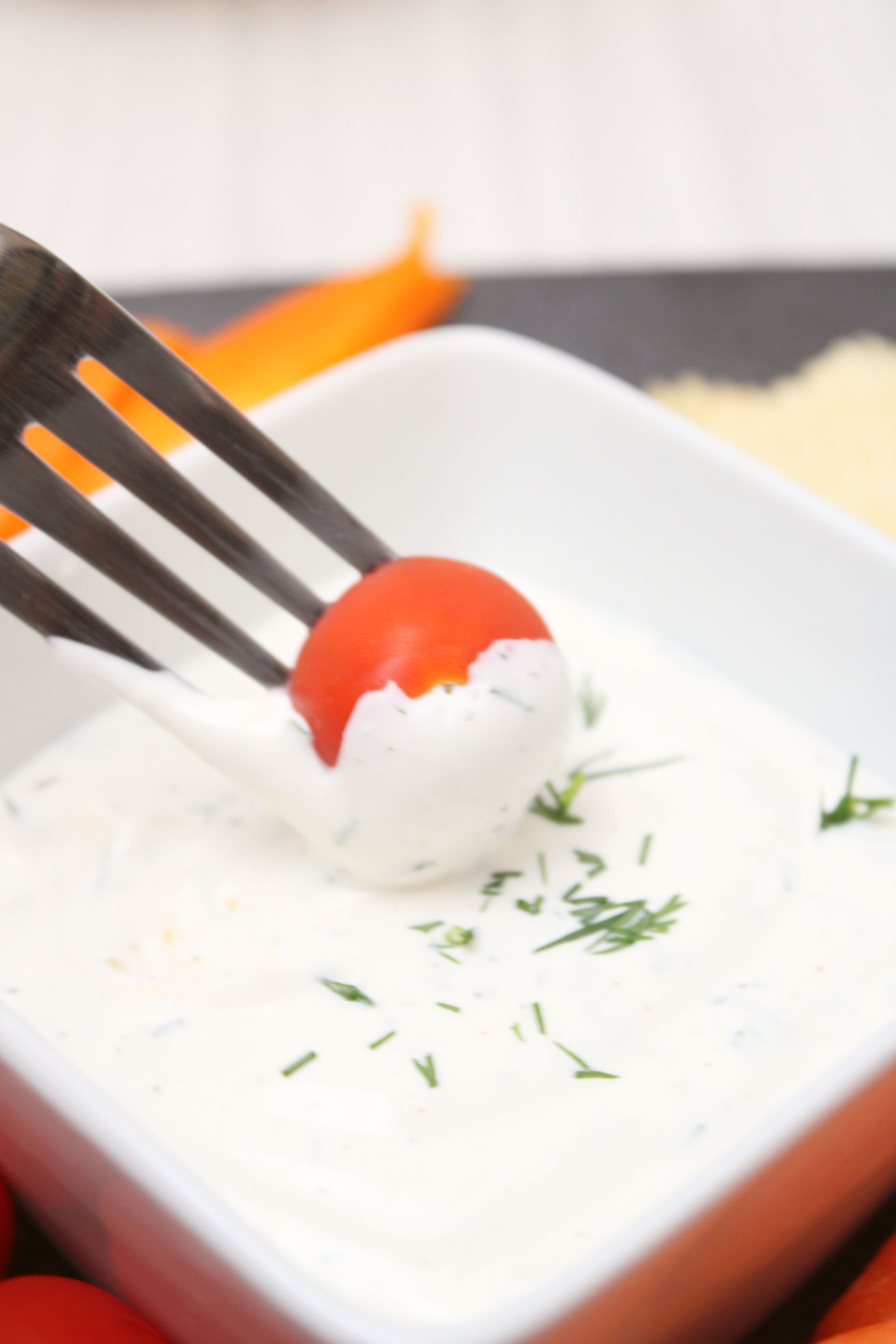 a cherry tomato on a fork being dipped into a white dip topped with fresh herbs