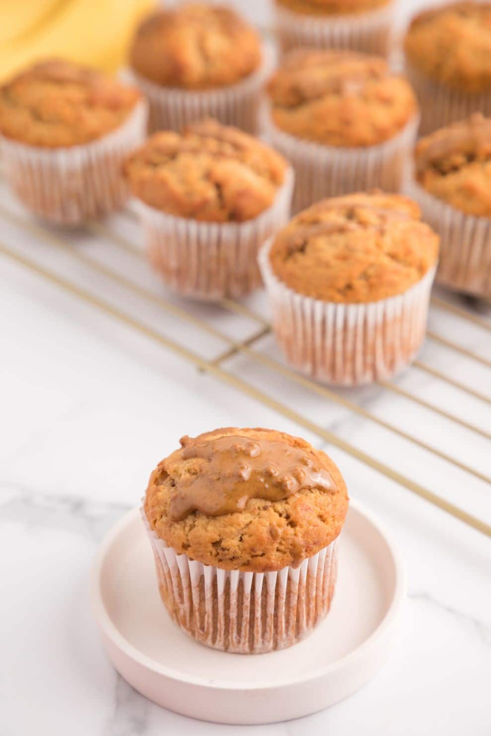 Baked muffins on a cooling rack with a small white plate containing one muffin topped with a Biscoff drizzle