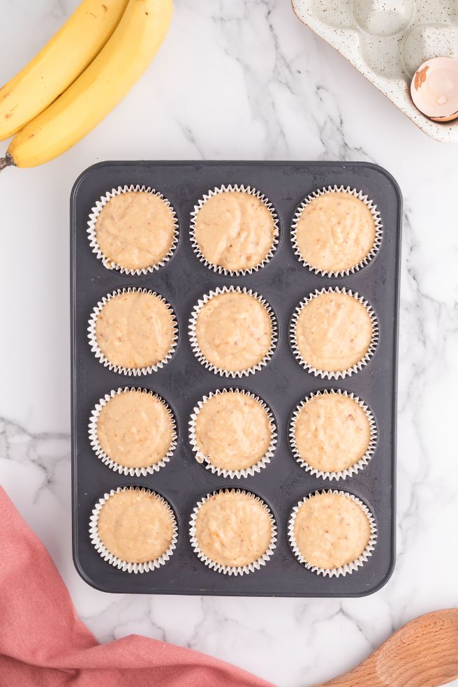 A 12 hole muffin pan containing muffin batter with some bananas in the background