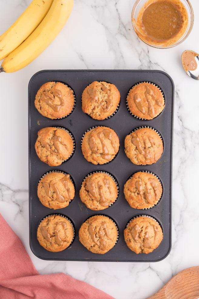 Baked muffins in a 12 hole muffin pan with some bananas and a Biscoff spread in the background