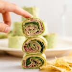 A stack of 3 spinach pinwheel sandwiches one on top of the other with a finger holding them together on top