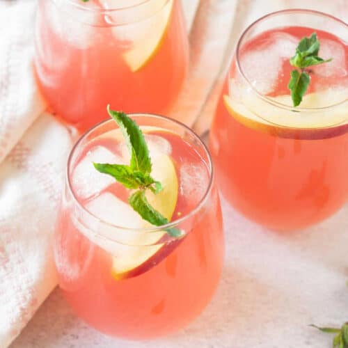 pink lemonade served in 3 glasses along with slices of fresh peach and sprigs of mint